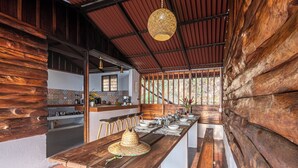A wooden interior with a long dining table set for a meal, a kitchen area, and a view through large windows. - Nebula Minca Ecolodge by Xarm Apartments (Santa Marta)