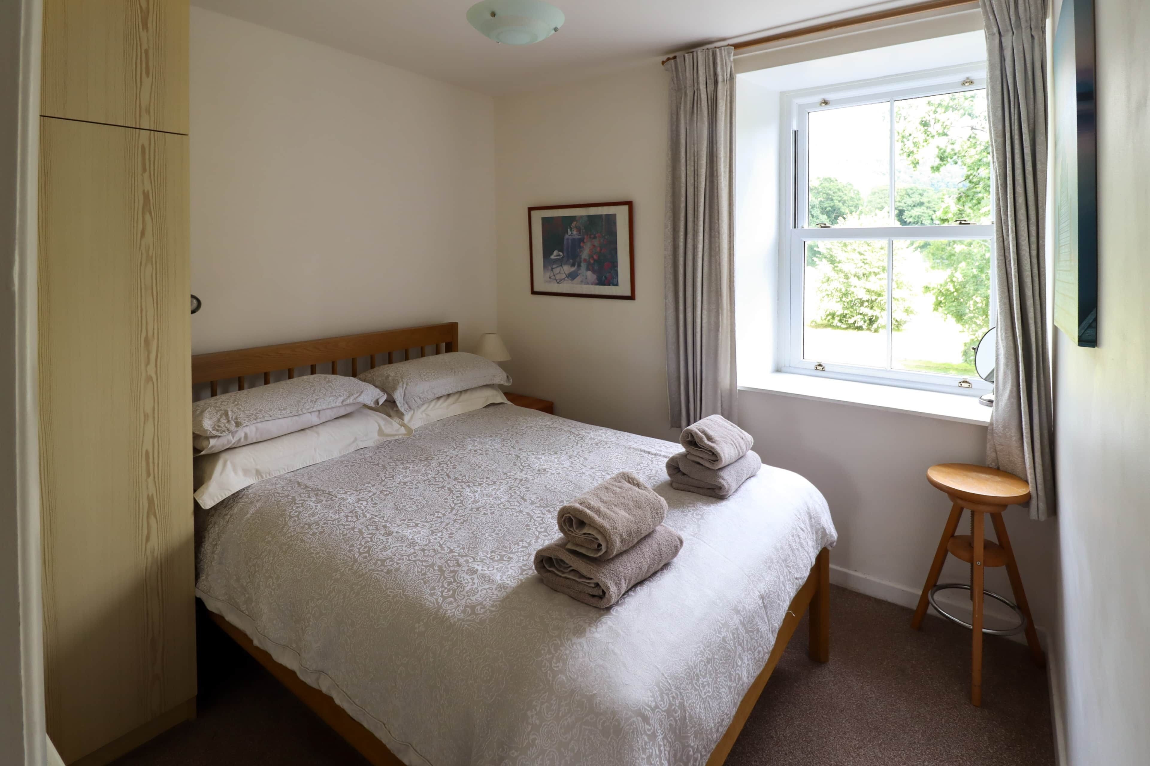 A bedroom with a bed, a window with curtains, a wooden stool, and a framed picture on the wall.