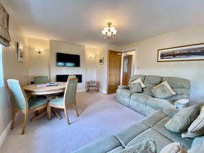 A living room with a sofa, chairs, a dining table, a TV, and a door. - 8 Howrahs Court, Elliott Park, Keswick (Keswick)