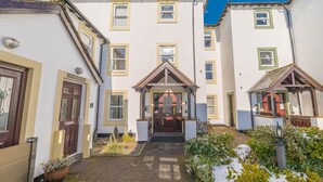 A building with a central entrance, multiple windows, and a small garden area with plants and a lamp post. - Riverside View, 5 Elm Court, Elliott Park (Keswick)
