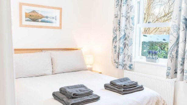 A neatly made bed with pillows, a bedside lamp, and a window with curtains. - Riverside View, 5 Elm Court, Elliott Park (Keswick)