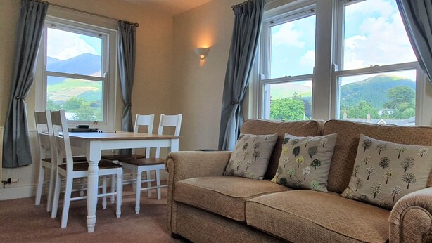 A room with a brown sofa, white chairs, a table, and large windows with curtains. - Skiddaw, 20 Greta Grove House (Keswick)