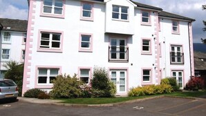 A multi-story residential building with pink-framed windows, a central balcony, and a parking lot in front. - Skiddaw, 20 Greta Grove House (Keswick)