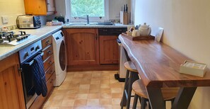 A kitchen with wooden cabinets, a built-in dishwasher, a stove, a washing machine, and a window providing natural light. - Skiddaw, 20 Greta Grove House (Keswick)