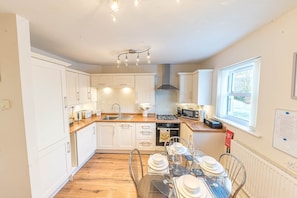 A modern kitchen with white cabinets, a central island, and a dining area with a table set for four. - Lilac Cottage, 17 Elm Court, Keswick (Keswick)