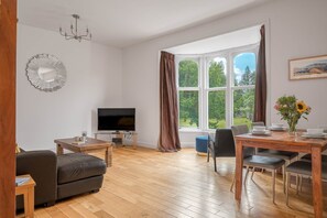 A living room with a TV, wooden furniture, a dining table, and a large window with curtains.