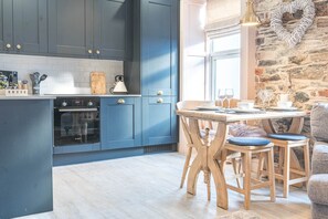 A modern kitchen with dark blue cabinets, a built-in oven, and a dining area with wooden chairs and a table set for two. - Withesike, Apartment 2, Derwent House (Portinscale)
