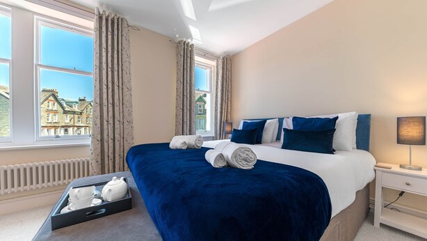 Une chambre d’hôtel avec un grand lit, une table de chevet et un plateau avec un service à thé, donnant sur les bâtiments environnants.