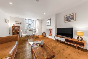 A living room with a sofa, coffee table, TV, bookshelf, and a dining area with chairs and a table. - Chaucer Retreat, 3 Chaucer House Apartments (Keswick)