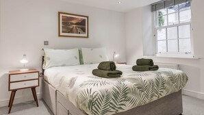 Une chambre à coucher avec un lit, une table de chevet, une lampe, un tableau encadré au mur et une fenêtre avec des stores.