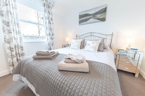 A neatly made bed with a quilted bedspread, two bedside lamps, a bedside table with a lamp, and a window with curtains. - Felltops, 6 Lonsdale House, Elliott Park (Keswick)