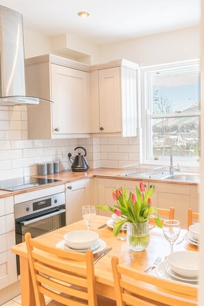 A modern kitchen with white cabinets, a stainless steel range hood, a kettle, and a dining area with a table set for two. - 7 Greta Grove House, Elliott Park (Keswick)