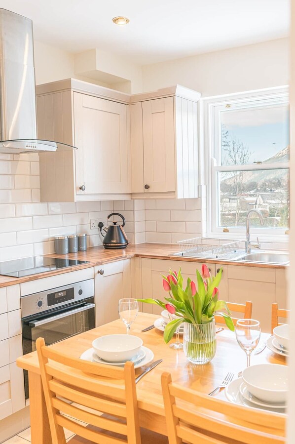 A modern kitchen with white cabinets, a stainless steel range hood, a kettle, and a dining area with a table set for two. - 7 Greta Grove House, Elliott Park (Keswick)