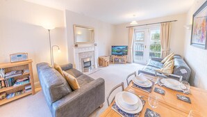 A living room with a sofa, a fireplace, a television, and a dining area with a table set for two. - 16 Greta Grove House (Keswick)