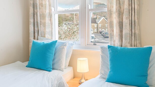A bedroom with two beds, a nightstand with a lamp, and a window with curtains. - 16 Greta Grove House (Keswick)