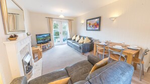 A living room with a fireplace, a sofa, a dining table with chairs, a television, and a balcony with curtains. - 16 Greta Grove House (Keswick)