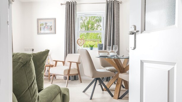 Een kamer met een groene bank, een eettafel met stoelen, een raam met gordijnen en een deur.