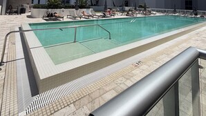 Una piscina con plataforma de baldosas, palmeras y edificios altos al fondo.