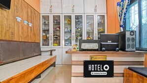 A reception desk with a sign indicating 'HOTEL' and 'Company Serviced.' There are shoe cabinets in the background, a bench, and a window with curtains. - Deluxe Ranihati Tripureswari (Howrah)