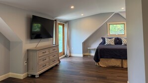 A bedroom with a TV mounted on the wall, a bed with patterned pillows, a dresser, and a window with a view of greenery. - BRAND NEW, Complete renovation: 4-Bedroom, 3.5 Bath, private Annisquam Gem! (Gloucester)