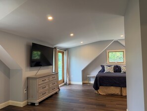 Ein Schlafzimmer mit an der Wand montiertem Fernseher, einem Bett mit gemusterten Kissen, einem Schminktisch und einem Fenster mit Blick ins Grüne.