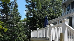A wooden deck with a white railing, surrounded by a yard with a lawn mower and trees. - BRAND NEW, Complete renovation: 4-Bedroom, 3.5 Bath, private Annisquam Gem! (Gloucester)