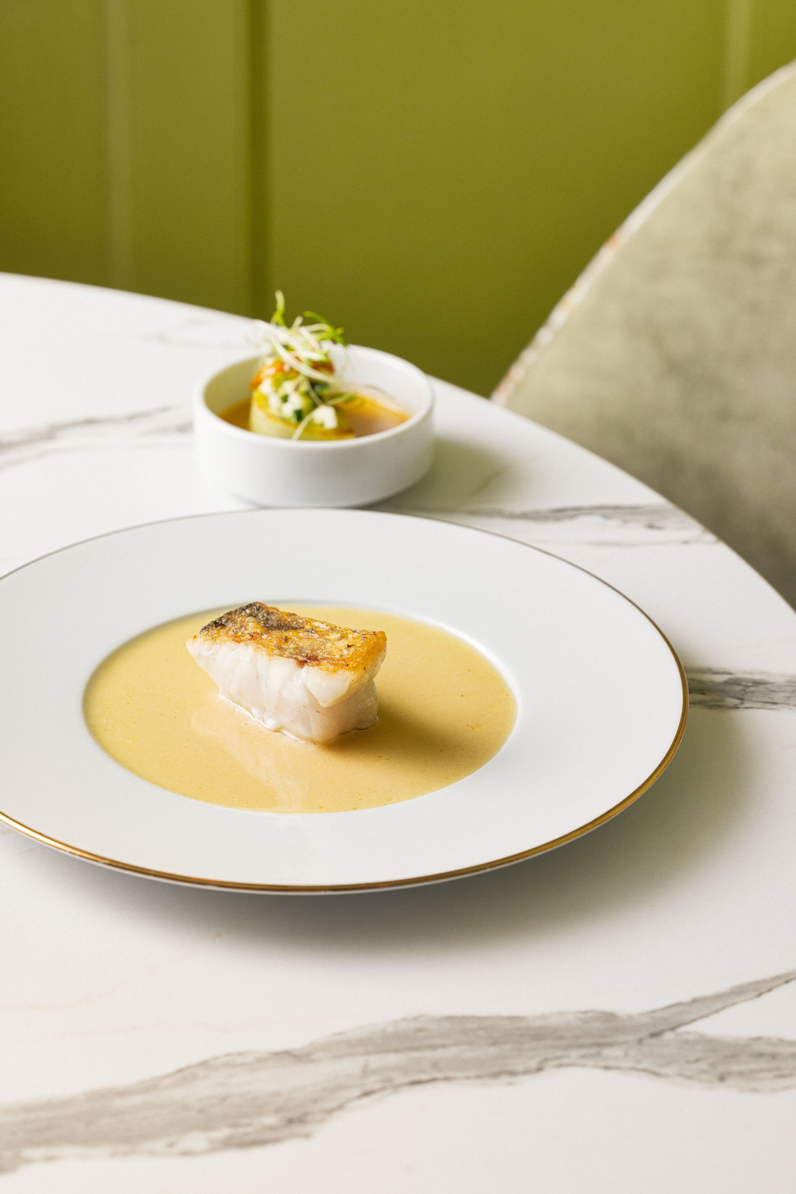 A plate of food with a piece of fish on a white plate with a gold rim, accompanied by a small bowl of salad and sauce.