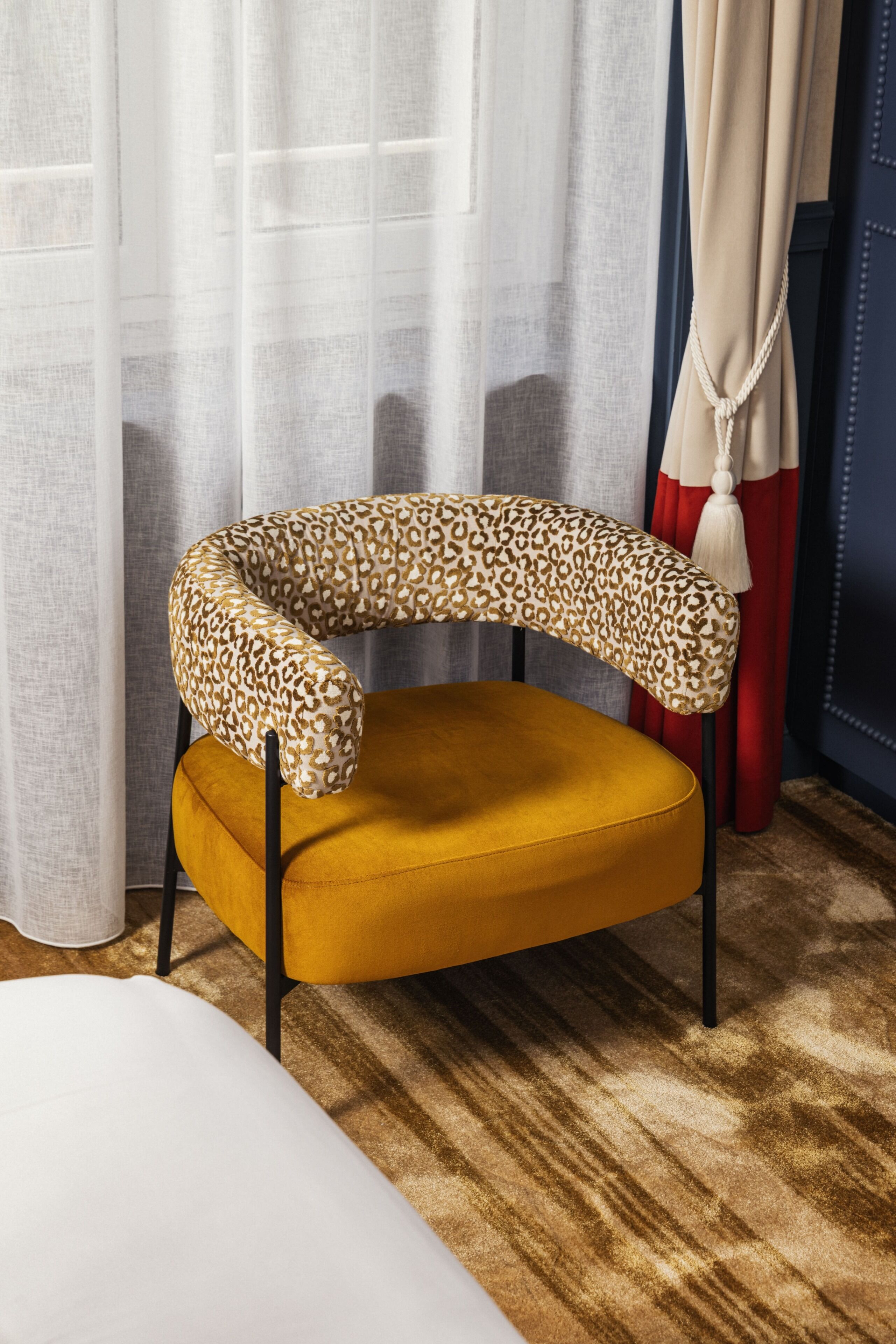 A modern chair with a unique curved backrest and a solid seat, positioned in a room with a patterned carpet and a window with sheer curtains.