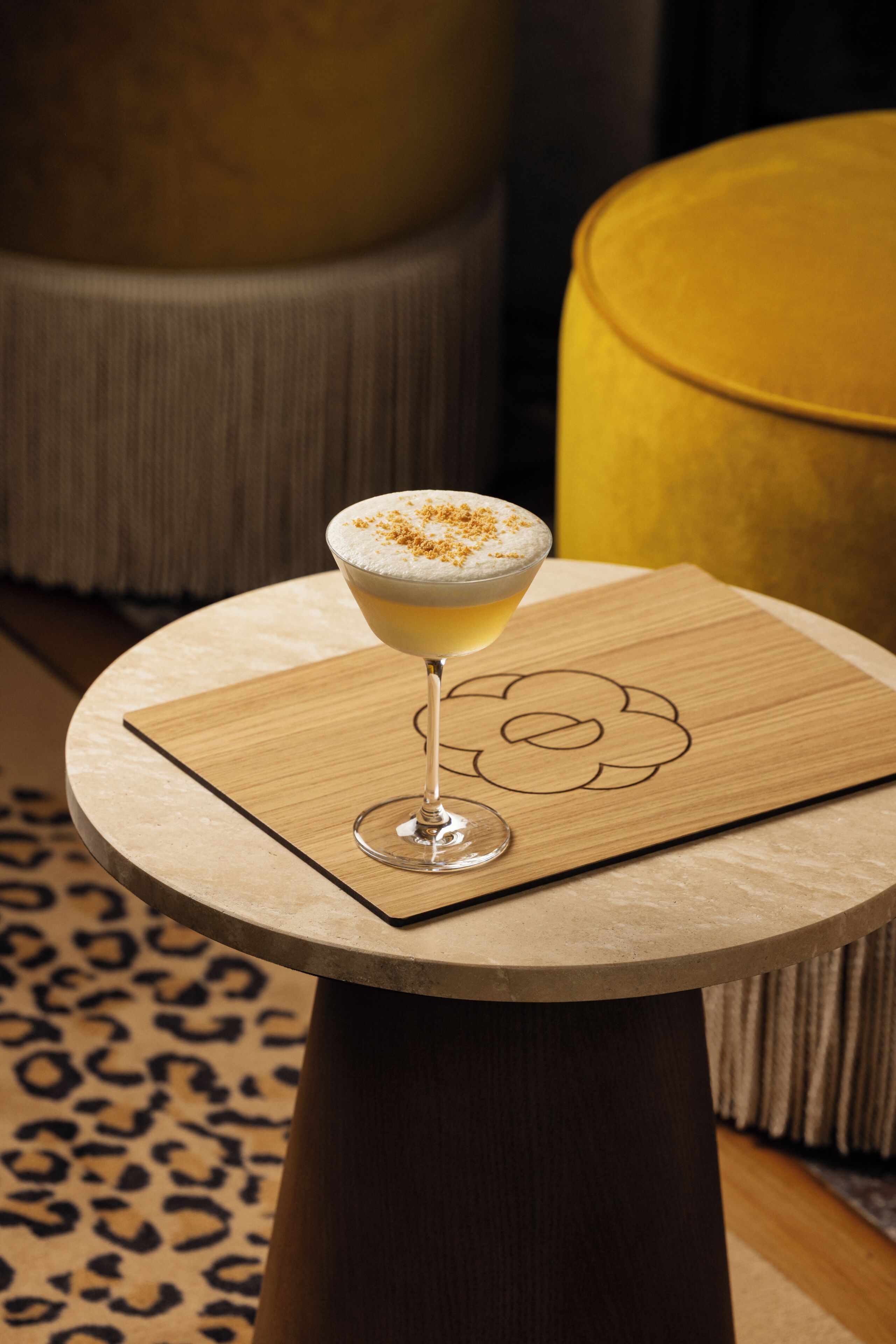 A cocktail on a round table with a wooden coaster, a patterned rug, and a yellow ottoman in the background.