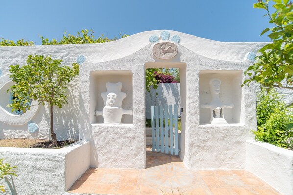 Une entrée blanche, à la texture particulière, agrémentée d’un portail, d’un petit arbre et d’un emblème décoratif placé au-dessus du portail.
