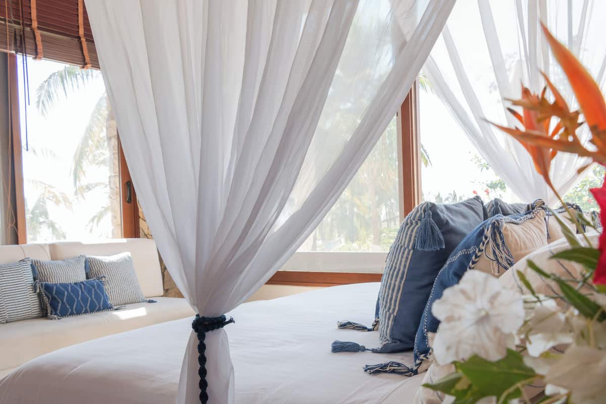 Ein Bett mit Baldachin, eine Couch mit gemusterten Kissen und Blick auf Palmen durch große Fenster.