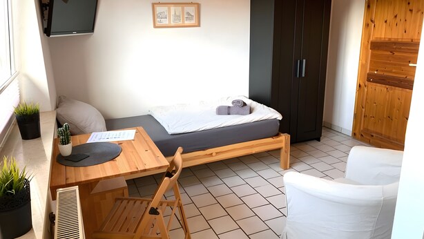 Ein kleiner, ordentlicher Raum mit einem Bett, einem Holztisch, einem Stuhl und einem Kleiderschrank.