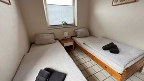 Une petite chambre propre, avec deux lits simples, une table de chevet, une fenêtre avec des stores et une plante en pot.