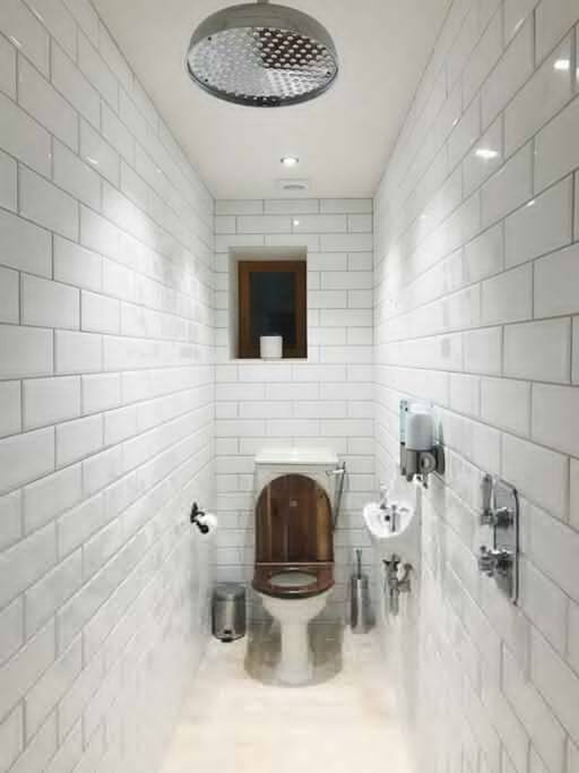 A narrow bathroom with white tiled walls, a wooden toilet, a wall-mounted sink, and a ceiling light fixture.