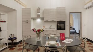 Een eethoek met een ronde tafel, stoelen, een bloevas, een koffiepot en een rood bord aan de muur.