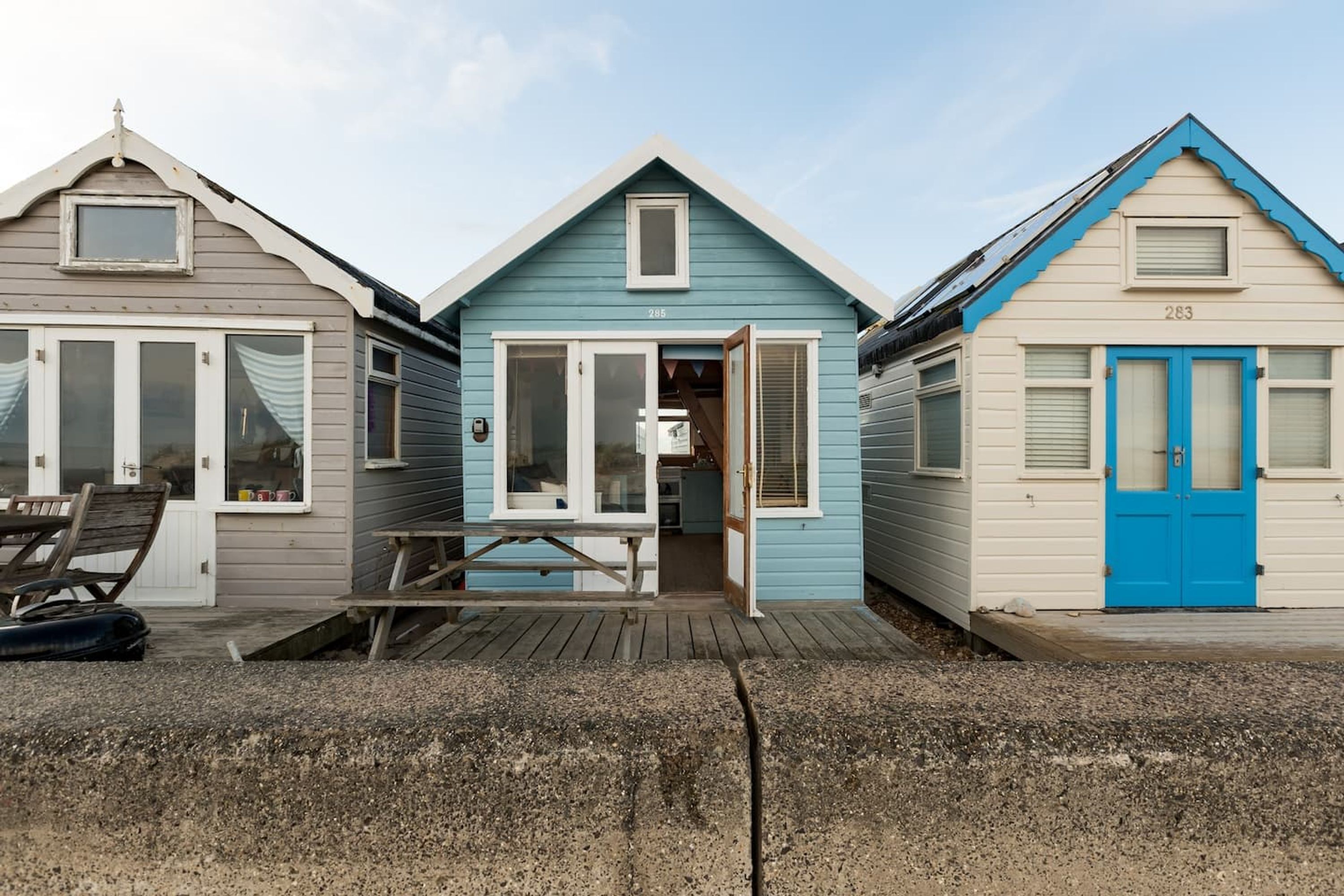 Three beach houses with distinct colors and numbers, a wooden deck, and a bench.