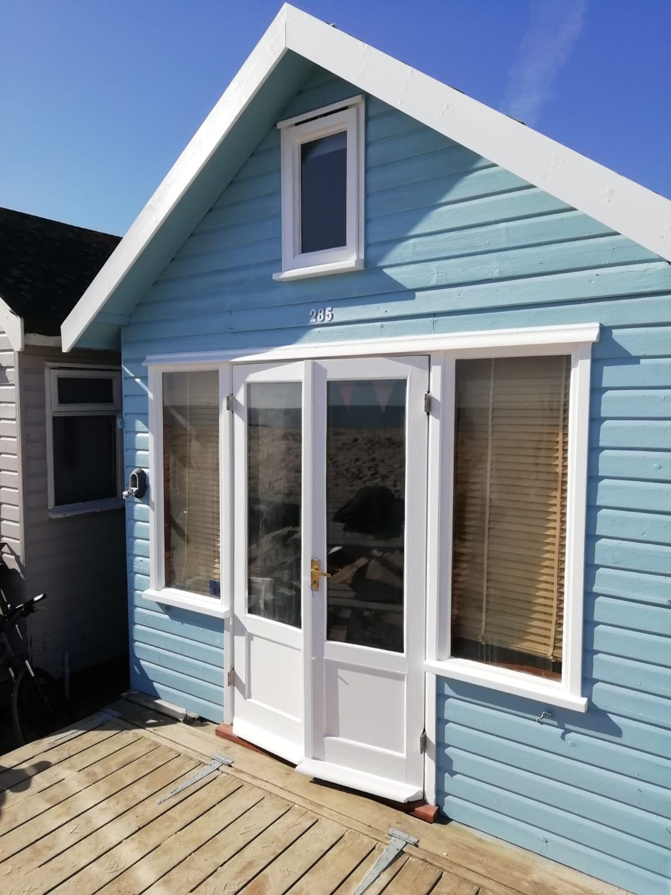 A light blue building with a white door and windows, number 285, and a wooden deck in front.