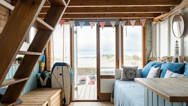 A wooden interior with a bed, a table, and a view of the beach. - Stargaze From a Beach hut on the Mudeford Sandbank (Bournemouth)