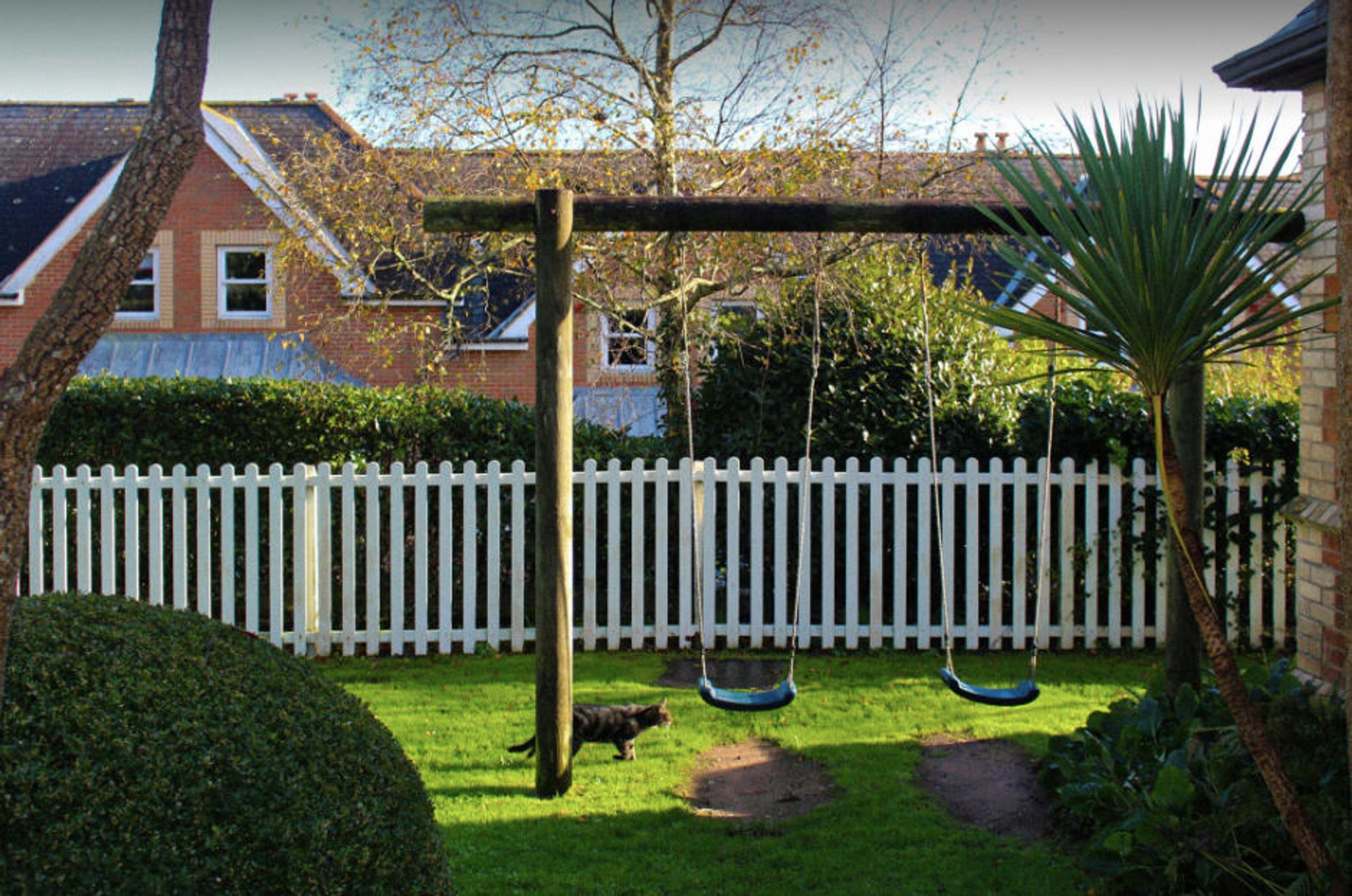 A backyard with a swing set, a white picket fence, and a house in the background.