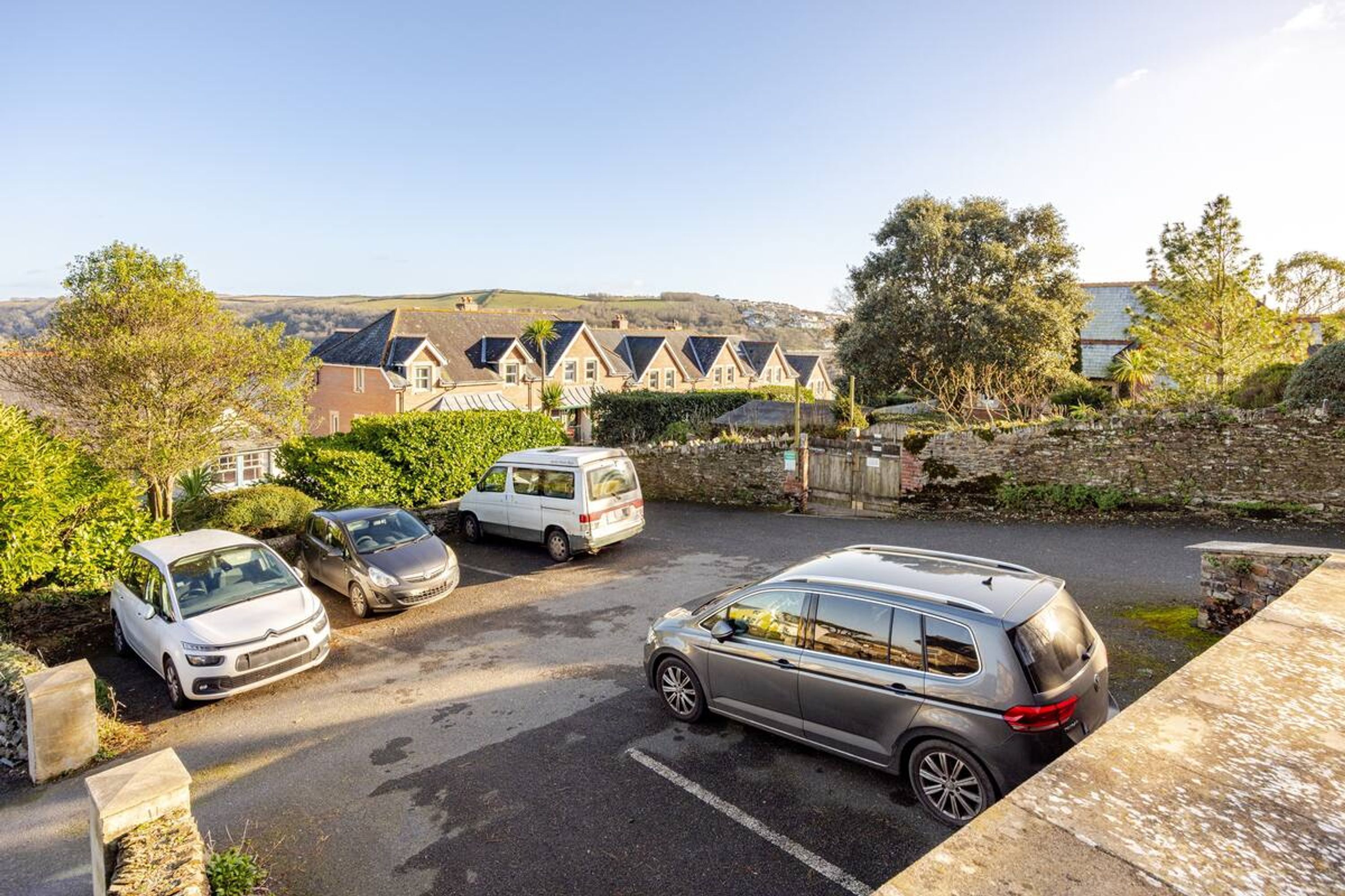 A parking area with cars and a van, surrounded by greenery and a stone wall.