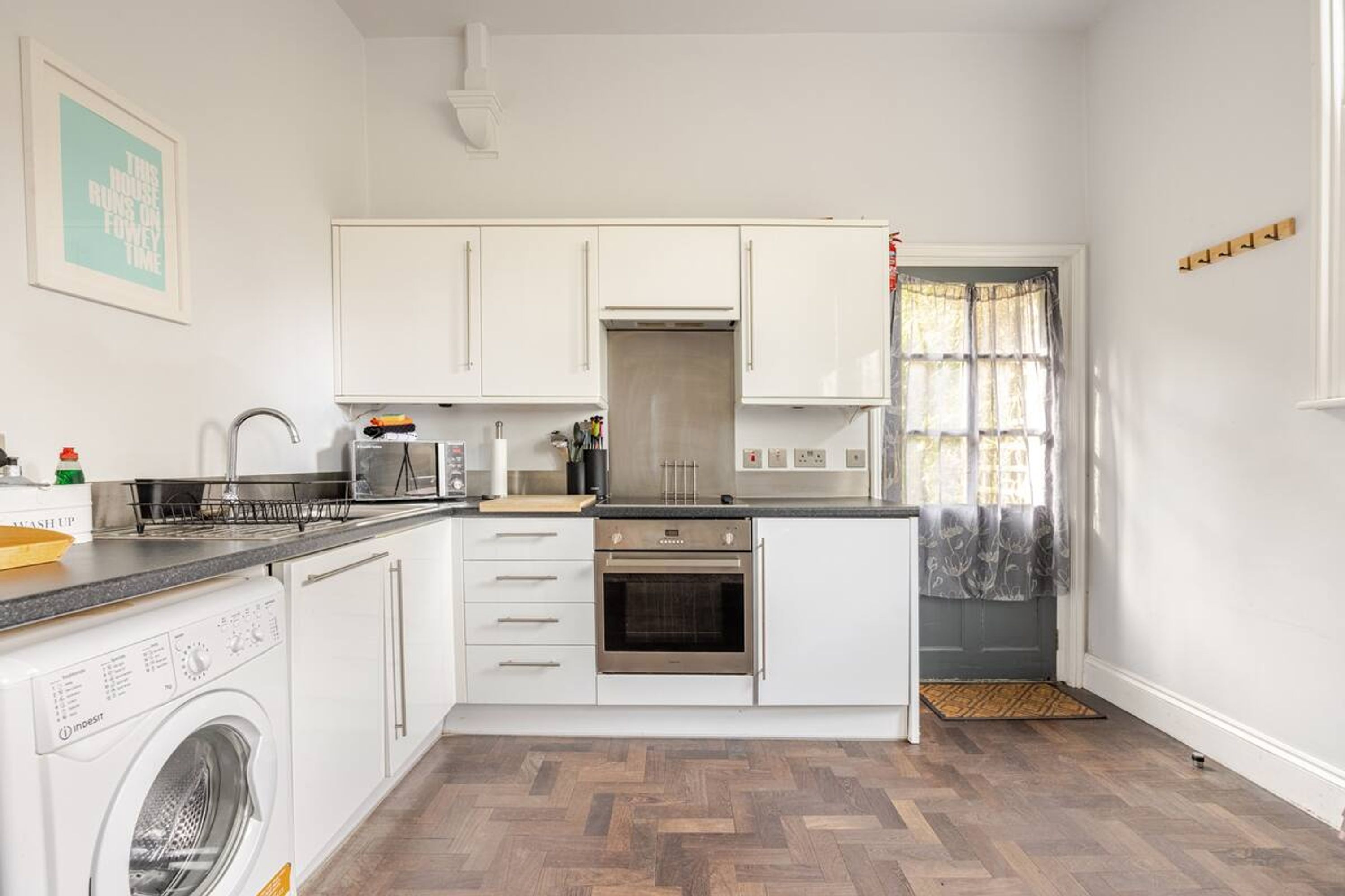Une cuisine compacte avec des meubles blancs, un four encastré, un lave-linge et une fenêtre avec des rideaux.