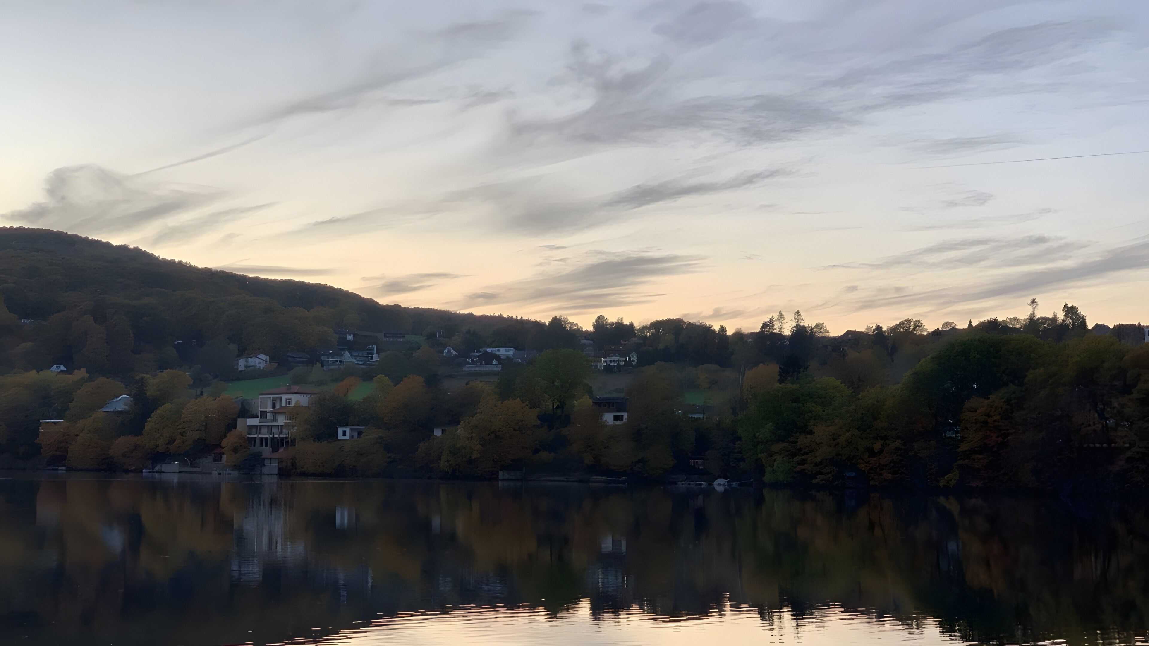 En stilla sjö där träd och hus speglas i vattnet, omgiven av kullar och en klar himmel.