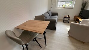 A modern living room with a wooden table, two grey armchairs, a grey sofa, a small side table with a plant, and a TV on a wooden stand. - Apartment for 2 People (25 m²) in Kreuzau (Kreuzau)