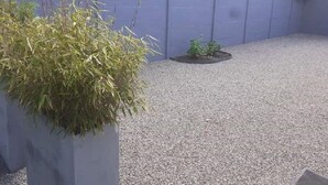 Uma área de pátio com calçamento de cascalho, um pequeno canteiro de plantas e um vaso alto.
