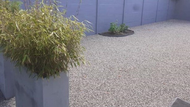 Un área de patio con grava, un pequeño macizo de plantas y una planta alta en maceta.