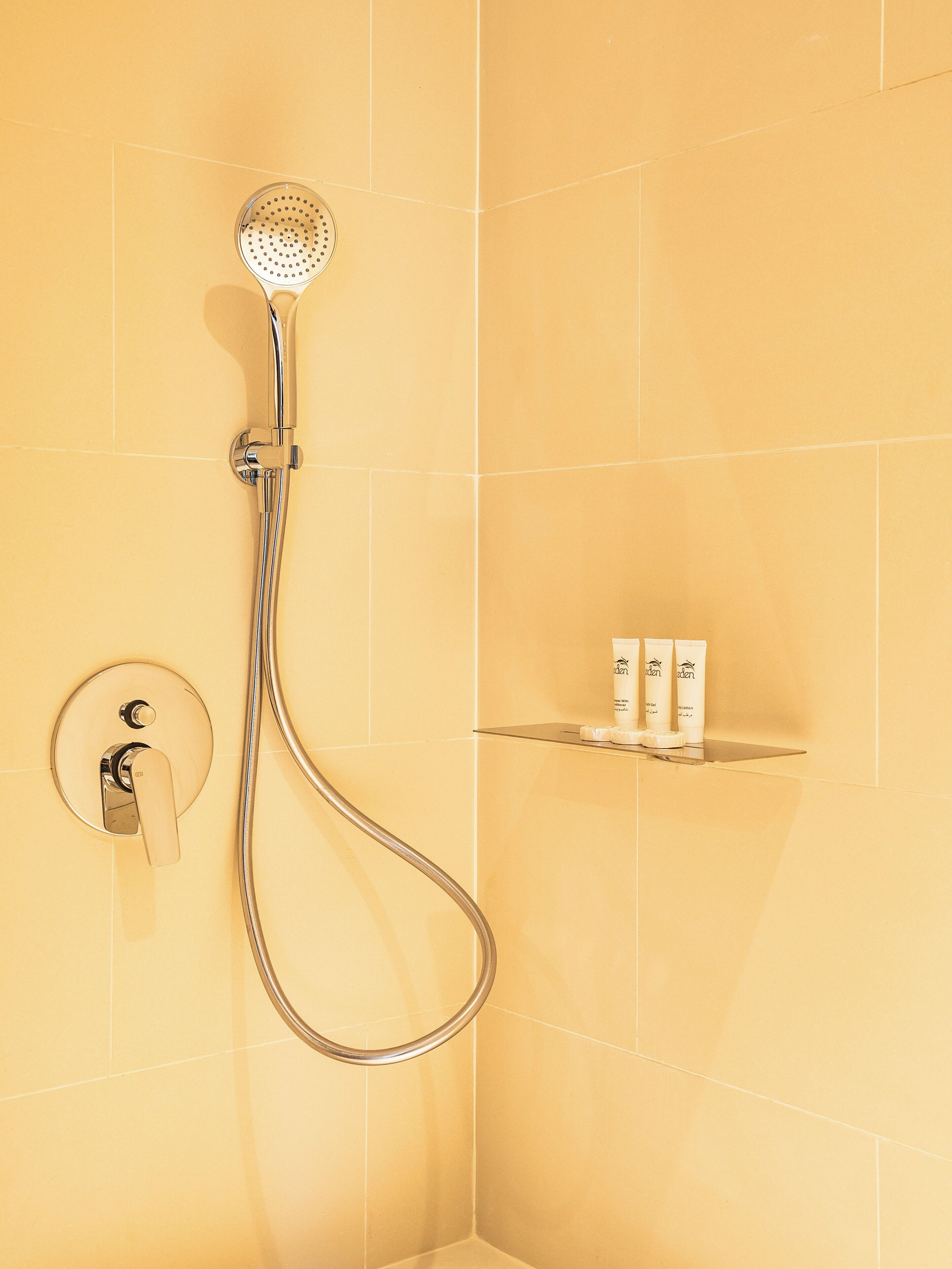 A shower with a handheld showerhead, a wall-mounted control, and a shelf with toiletries.