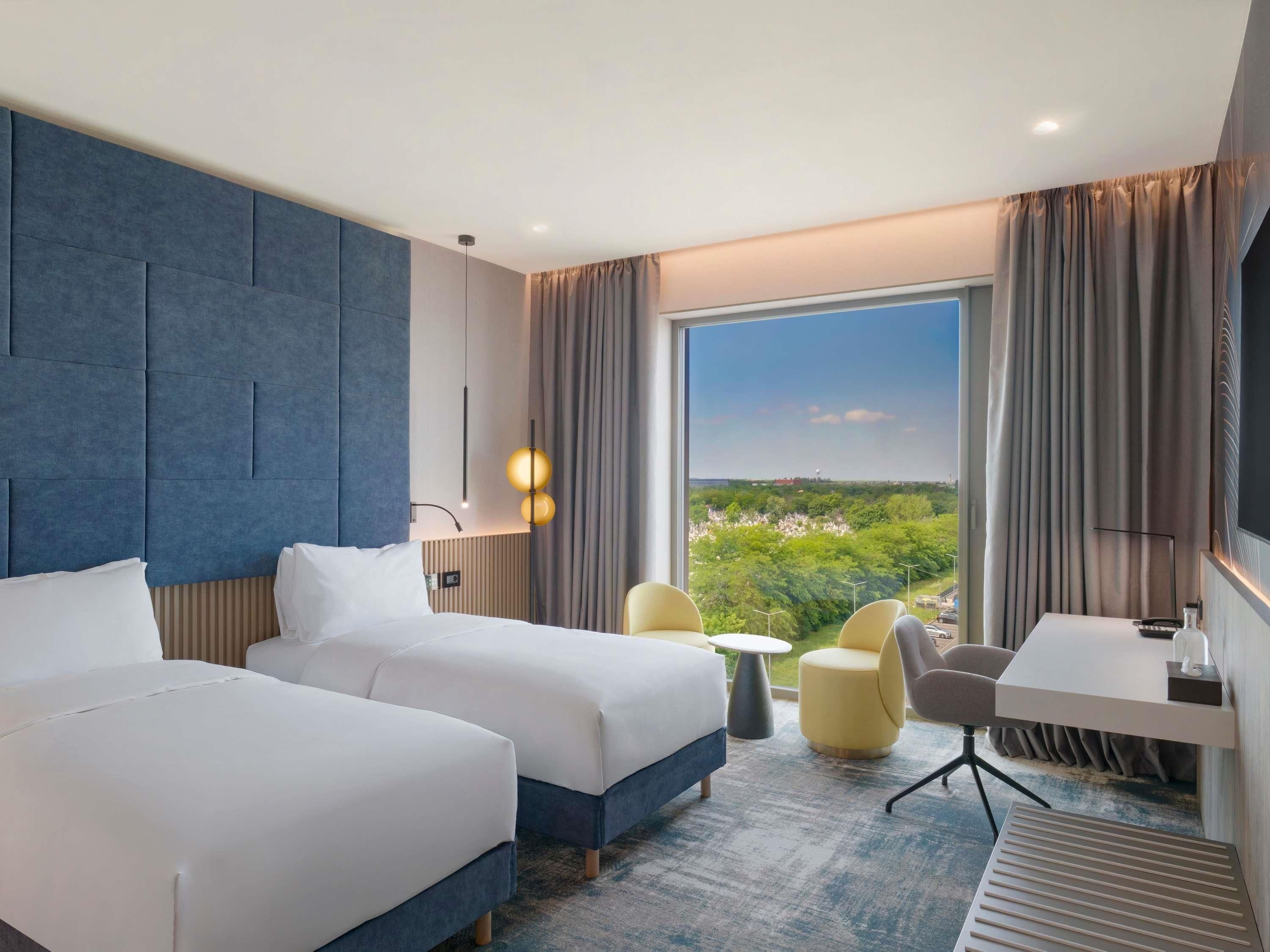 Een hotelkamer met twee bedden, een bureau, een stoel, een klein tafeltje en een groot raam met uitzicht op groen en gebouwen.