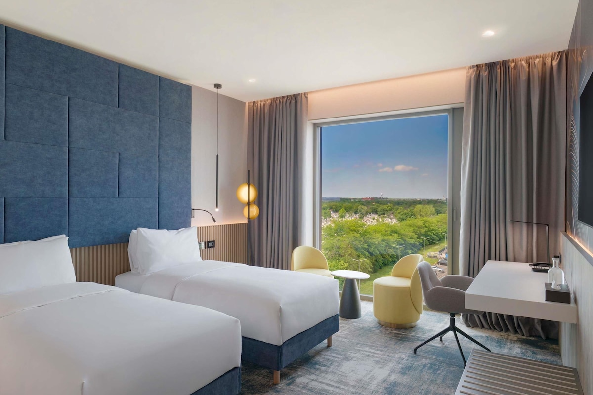Habitación de hotel con dos camas, un escritorio, una silla, una mesita y un ventanal grande con vistas a zonas verdes y edificios.