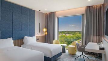 Een hotelkamer met twee bedden, een bureau, een stoel, een klein tafeltje en een groot raam met uitzicht op groen en gebouwen.