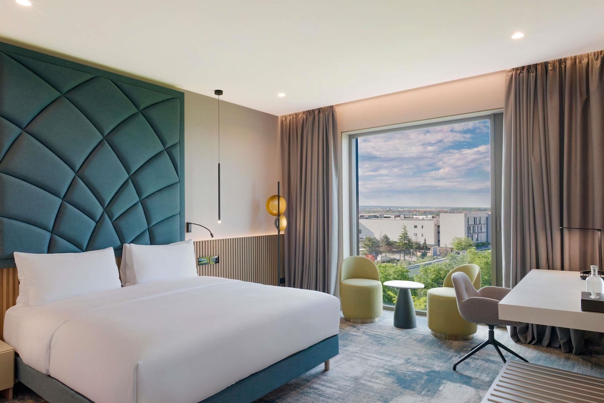 Una habitación de hotel moderna con una cama grande, un escritorio, dos sillas y vista a la ciudad.